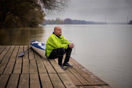 Photo for A middle-aged man sitting on the jetty and ready to go kayaking. Active sporty man wearing dry suit and looking away. - Royalty Free Image