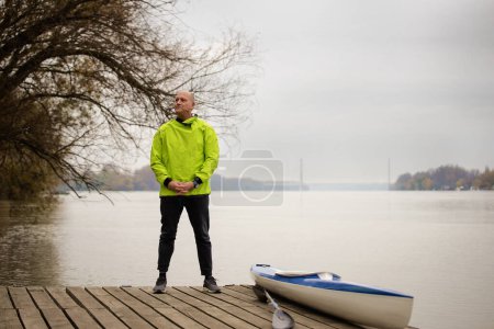Téléchargez les photos : Un homme d'âge moyen debout sur la jetée et prêt à faire du kayak. Homme sportif actif portant une combinaison sèche. Plan complet. - en image libre de droit