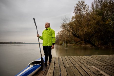 Photo for A middle-aged man standing on the jetty and ready to go kayaking. Active sporty man wearing dry suit and looking away and holding oar in his hand. - Royalty Free Image