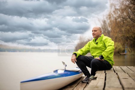 Foto de Un hombre de mediana edad sentado en el embarcadero y listo para ir en kayak. Hombre deportivo activo con traje seco y mirando hacia otro lado. - Imagen libre de derechos