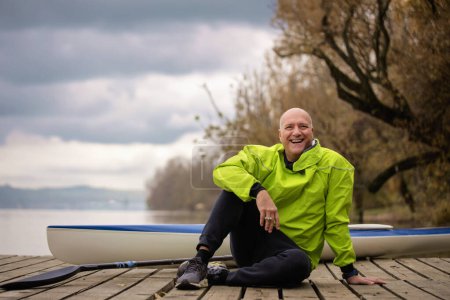 Photo for A middle-aged man sitting on the jetty and ready to go kayaking. Active sporty man wearing dry suit and looking at camera. - Royalty Free Image