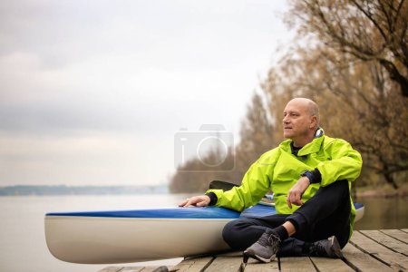 Téléchargez les photos : Un homme d'âge moyen assis sur la jetée et prêt à faire du kayak. Homme sportif actif portant une combinaison sèche et détournant les yeux. - en image libre de droit