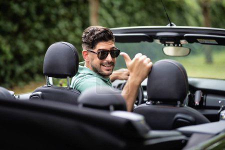 Photo for A handsome man wearing sunglasses and sitting in a convertible car and driving on the road. Happy caucasian male showing thumbs up. - Royalty Free Image