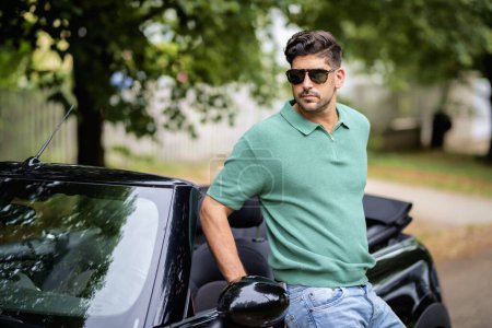 Foto de Un hombre guapo con gafas de sol y de pie junto a su coche convertible en la carretera. - Imagen libre de derechos