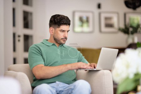 Téléchargez les photos : Homme d'âge moyen assis dans son fauteuil roulant à la maison et utilisant un ordinateur portable pour le travail. Homme d'affaires confiant portant des vêtements décontractés. Bureau à domicile. - en image libre de droit