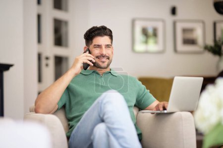 Photo for Mid aged man sitting at home and using laptop and smartphone. Confident male making a call. Home office. - Royalty Free Image