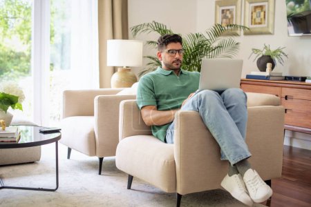 Photo for Mid aged man sitting in his amrchair at home and using laptop for work or having video call. Confident businessman wearing casual clothes. Home office. Full length shot. - Royalty Free Image