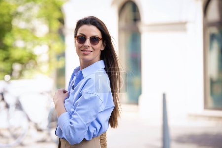 Foto de Mujer morena feliz con gafas de sol y camisa azul y caminando por la calle en la ciudad. - Imagen libre de derechos