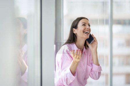 Photo for Confident businesswoman standing by the window in a modern office and making a call. Brunette haired woman wearing business casual. - Royalty Free Image