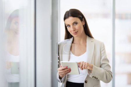 Téléchargez les photos : Femme d'affaires confiante debout dans un bureau moderne et utilisant un pavé tactile pour le travail. Femme attrayante portant un blazer et regardant la caméra. - en image libre de droit