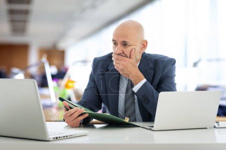 Téléchargez les photos : Homme d'affaires d'âge moyen assis dans le bureau et utilisant un ordinateur portable pour le travail. Homme professionnel confiant portant costume et cravate et regardant le carnet soigneusement. - en image libre de droit