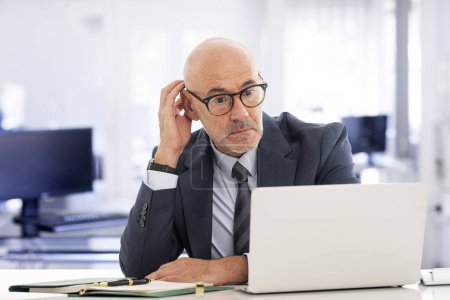 Téléchargez les photos : Homme d'affaires d'âge moyen assis dans le bureau et utilisant un ordinateur portable pour le travail. Homme professionnel confiant portant costume et cravate et regardant le carnet avec soin. Il fait une vidéoconférence.. - en image libre de droit