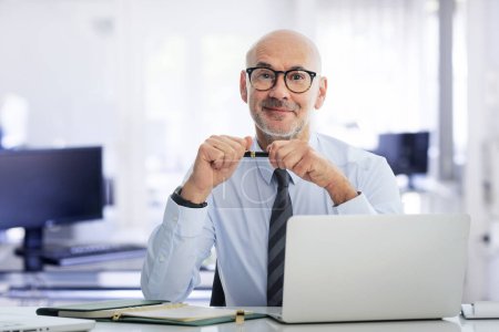 Téléchargez les photos : Homme d'affaires d'âge moyen assis dans le bureau et utilisant un ordinateur portable pour le travail. Homme professionnel confiant portant chemise et cravate. - en image libre de droit