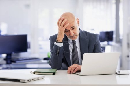 Téléchargez les photos : Un homme d'affaires d'âge moyen assis à son bureau et utilisant un ordinateur portable. Il regarde l'ordinateur portable avec attention. Homme professionnel exécutif ayant une vidéoconférence. - en image libre de droit