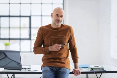 Foto de Un hombre de mediana edad de pie en su escritorio en la oficina y mirando reflexivo. Hombre seguro usando ropa casual. Copiar espacio. - Imagen libre de derechos