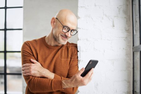 Photo for Middle-aged man standing at the window at using smartphone. Smiling male wearing casual clothes and text messaging. - Royalty Free Image