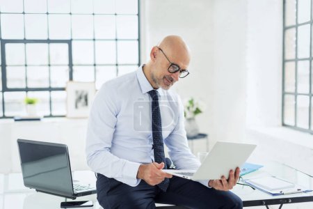 Téléchargez les photos : Un homme d'affaires debout dans le bureau avec un ordinateur portable à la main. Homme professionnel d'âge moyen portant costume et cravate. - en image libre de droit