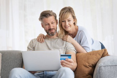 Téléchargez les photos : Achats en ligne de la maison. Beau homme tenant la carte bancaire dans sa main tandis que sa femme assise à côté de lui et en utilisant un ordinateur portable tout en faisant des achats en ligne. - en image libre de droit