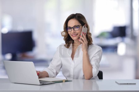 Téléchargez les photos : Joyeux sourire femme d'affaires d'âge moyen ayant un appel et en utilisant un ordinateur portable au bureau. - en image libre de droit