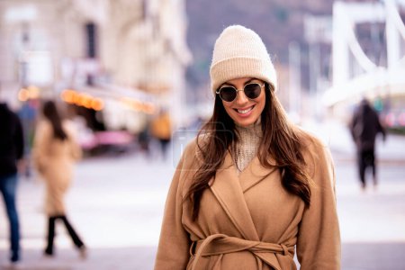Téléchargez les photos : Une femme d'âge moyen aux cheveux bruns se promène dans la rue du centre-ville. Attractiv femme portant des lunettes de soleil et manteau d'hiver. - en image libre de droit