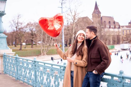 Téléchargez les photos : Couple debout ensemble en ville. Ils tiennent le ballon en forme de cœur dans la main et célèbrent la Saint-Valentin. Femme heureuse et bel homme célébrant leur amour - en image libre de droit