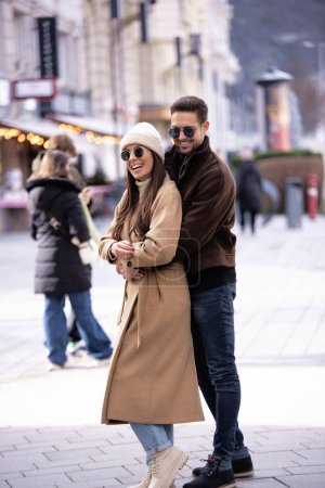 Foto de Longitud completa de una pareja feliz caminando por la calle y abrazándose. Morena mujer de pelo usando sombrero y abrigo y hombre guapo usando gafas de sol y abrigo. - Imagen libre de derechos