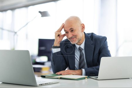 Téléchargez les photos : Homme d'affaires d'âge moyen assis dans le bureau et utilisant un ordinateur portable pour le travail. Homme professionnel confiant ayant une vidéoconférence. - en image libre de droit