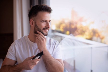 Shot of a man shaving his face with an electric shaver. He standing at the window and looking thoughtfully.