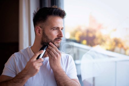 Shot of a man shaving his face with an electric shaver. He standing at the window and looking thoughtfully.