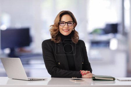 Téléchargez les photos : Femme d'affaires d'âge moyen assise au bureau et utilisant un ordinateur portable pour le travail. Femme professionnelle confiante portant une veste noire. Espace de copie. - en image libre de droit