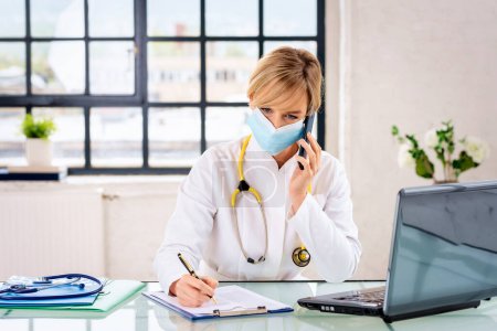 Photo for Female doctor sitting at the table and talking on the phone. Healthcare worker wearing surgical mask and using laptop. - Royalty Free Image