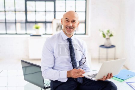 Téléchargez les photos : Homme d'affaires assis au bureau avec un ordinateur portable à la main. Homme professionnel d'âge moyen portant chemise et cravate et travaillant. - en image libre de droit