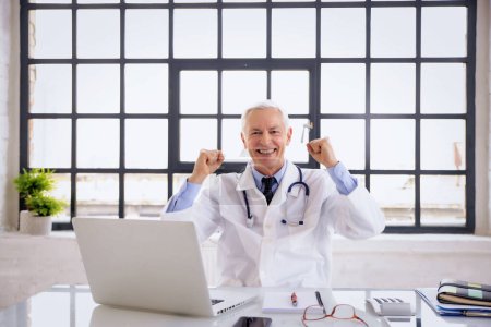 Porträt eines männlichen Arztes am Schreibtisch. Altenpfleger bedient Laptop und feiert seinen Erfolg. Er trägt Laborkittel.