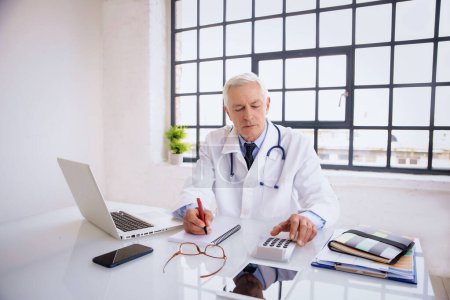 Portrait of male doctor sitting at desk. Elderly healthcare worker is working in hospital. He is wearing lab coat.