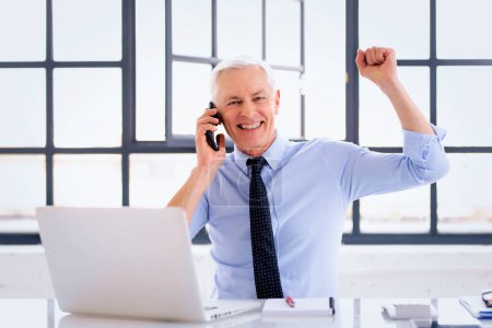 Foto de Hombre de negocios mayor sentado en su escritorio frente a su computadora y hablando en el teléfono inteligente. Levantando la mano y celebrando su éxito. - Imagen libre de derechos