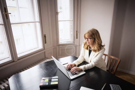 Foto de Una mujer de mediana edad está sentada en la mesa usando un portátil. Lleva una chaqueta blanca y se ve pensativa.. - Imagen libre de derechos