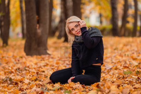 Photo for A girl with blond hair and glasses sits in an autumn park - Royalty Free Image