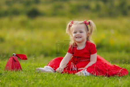 Photo for Little girl in a red dress sits on a green lawn - Royalty Free Image