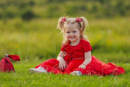 Photo for Little girl in a red dress sits on a green lawn - Royalty Free Image