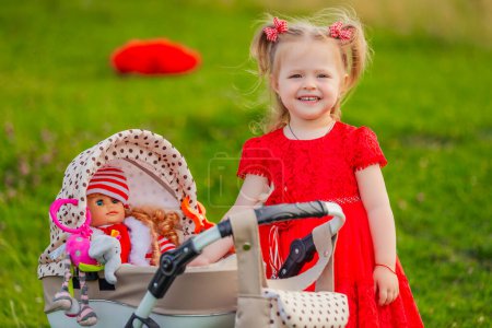 Photo for Little girl plays with a doll and a stroller on the lawn - Royalty Free Image