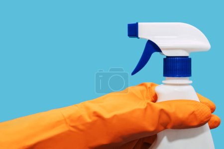 A human hand in a rubber orange glove holds a spray bottle with detergent on a blue background. The concept of cleaning and washing windows with detergent in a spray bottle