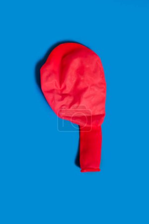 A deflated red balloon on a blue background. One uninflated balloon viewed from above. The concept of the ended holidays in the form of a half-deflated balloon