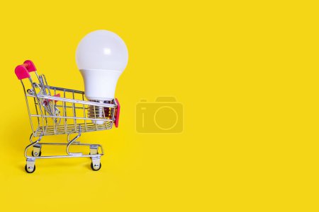 Photo for A small shopping cart and an energy-saving light bulb on a yellow background. Favorable purchase of electric power equipment to save costs. Sale of electrical equipment. Free space for text - Royalty Free Image