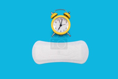 Foto de Almohadilla menstrual de higiene femenina para el ciclo menstrual y un despertador sobre fondo azul. El concepto de seguimiento del ciclo menstrual en forma de reloj y servilleta sanitaria - Imagen libre de derechos