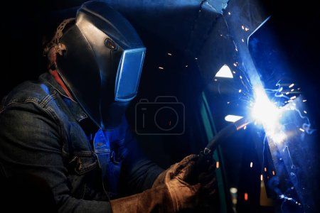 Un soldador trabaja usando equipo de soldadura y hace costuras en metal. El soldador lleva una máscara protectora y guantes. Chispas e incendios cuando se utiliza equipo de soldadura