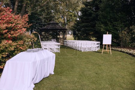 Foto de Evento al aire libre, sillas blancas colocadas fuera en el jardín, cenador de madera, caballete de madera con sábana blanca en blanco. Ceremonia de boda abierta en el jardín. Ceremonia de salida. Un lugar para celebrar. - Imagen libre de derechos