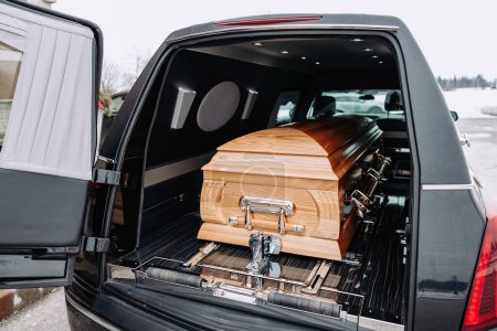 Photo for A coffin made of light wood stands in the trunk of a black hearse. Funeral and farewell ceremony. Closeup photo of a funeral casket. - Royalty Free Image
