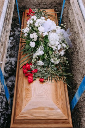 Photo for A wooden coffin in a grave. A coffin with white and red flowers on it is lowered into a dug grave. Funeral ceremony at the cemetery. Farewell ceremony and burial. Close-up photo. - Royalty Free Image