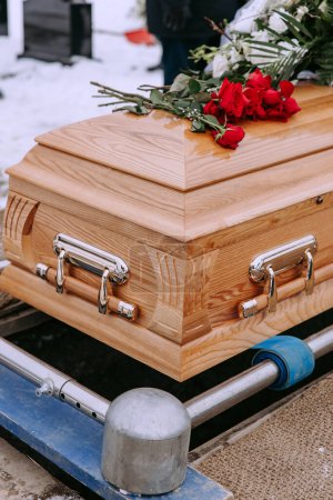 Un cercueil en bois avec des fleurs sur le couvercle se dresse au-dessus d'une tombe creusée dans le cimetière. Des roses rouges gisent sur le cercueil. Cérémonie funéraire au cimetière en hiver. Cérémonie d'adieu et enterrement. Photo en gros plan.