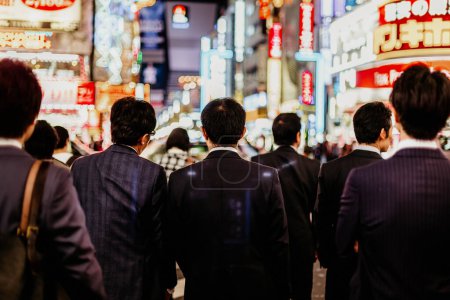 Foto de Empresarios corporativos japoneses en trajes, esperando en hora punta en la encrucijada en el distrito de negocios de Shinjuku, Tokio, Japón. Tableros publicitarios desenfocados iluminados en el fondo
. - Imagen libre de derechos